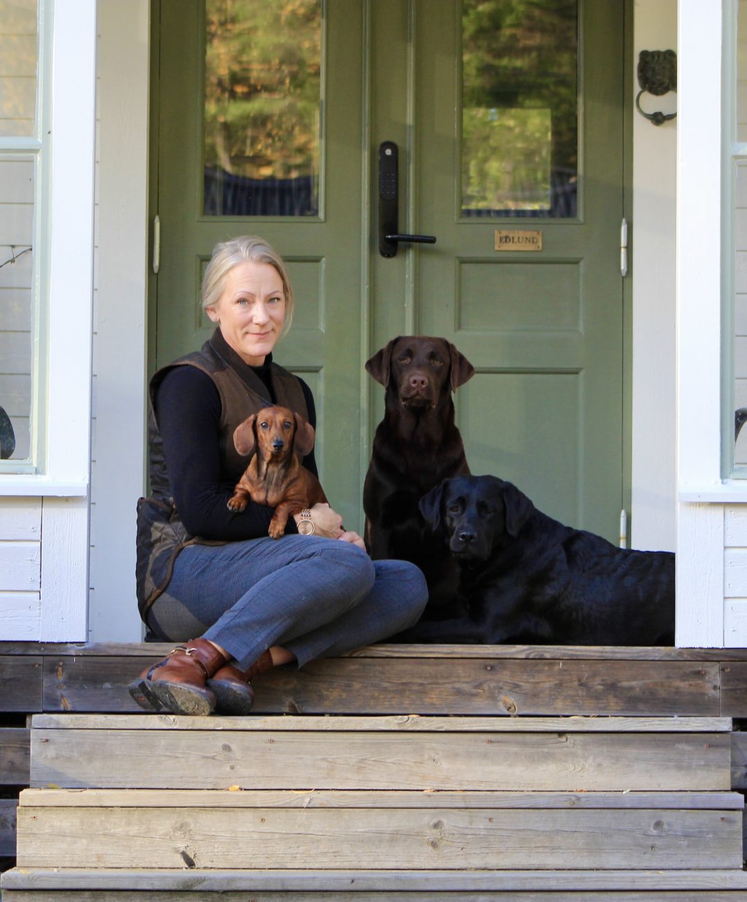 Louise Edlund on the front steps by the green door, with a dachshund in her lap and two labradors beside her